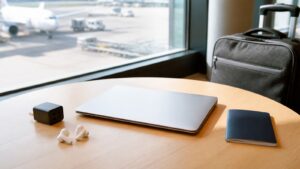 A closed lightweight laptop on an airport lounge table with travel accessories and a carry-on bag.